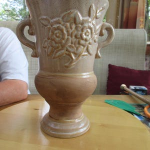 May include: A large, beige ceramic vase with two handles and a floral design. The vase has a textured surface and a wide base.