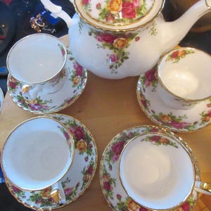 Peut inclure: Service à thé en porcelaine blanche comprenant une théière, des tasses et des soucoupes. L'ensemble est orné d'un motif floral de roses rouges, jaunes et roses, avec des bordures dorées. Les tasses sont posées sur des soucoupes, et la théière a un couvercle et un bec courbé.