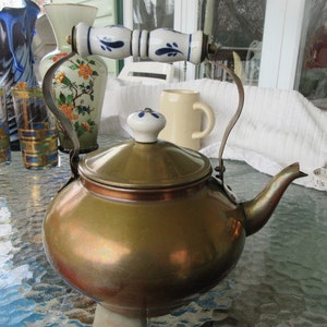 Peut inclure: Une bouilloire en cuivre vintage avec un couvercle et une poignée en porcelaine blanche. La poignée est décorée de motifs floraux bleus et blancs. La bouilloire est posée sur une table en verre.