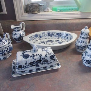 May include: A selection of white ceramic tableware featuring blue floral designs. The set comprises a sugar bowl, creamer jug, oval serving dish, butter dish, and two salt and pepper shakers. The items are displayed on a countertop.