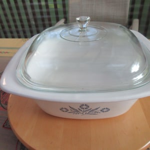 May include: White casserole dish with a clear glass lid. The dish has a square shape with rounded corners and a floral design in blue. The lid has a clear glass knob. The dish is sitting on a wooden surface.