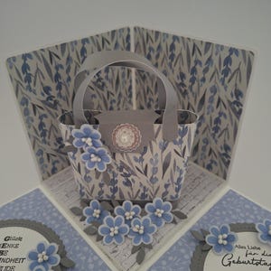 Könnte beinhalten: Handgefertigte Grußkarte mit einem Pop-up-Korb-Design. Der Korb ist mit blauen Blumenmustern verziert und mit Papierblumen geschmückt. Die Karte hat ein hellblaues und graues Farbschema, mit Text auf der Innenseite.