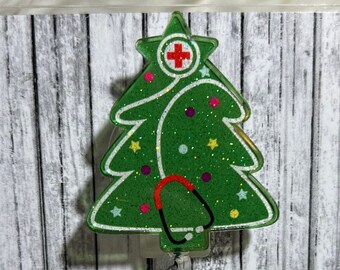 May include: A green glitter Christmas tree-shaped badge holder with a red cross and a stethoscope. The badge holder is on a white wood background.