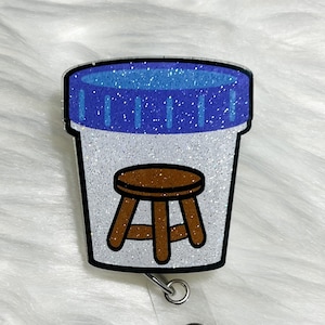 May include: A white glitter badge reel with a blue glitter lid and a brown glitter stool inside. The reel has a clear plastic strap and a silver metal clip.