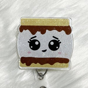 May include: A sparkly, light brown, white, and brown s'mores-themed badge reel with a cute cartoon face. The reel has a clear plastic backing and a clear plastic retractable cord.