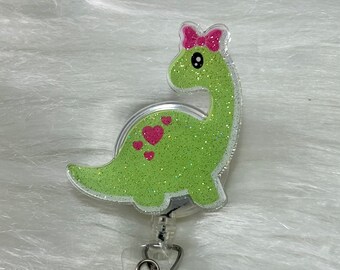 May include: Green glitter dinosaur with a pink bow and pink hearts on a clear plastic retractable badge holder with a silver clip and two silver swivel clips.