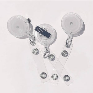 May include: Three clear plastic retractable badge reels with circular housings and metal belt clips. Each reel features a transparent vinyl strap attached by a silver metal ring, secured with a silver metal snap button for holding identification cards.