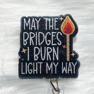 May include: A black glitter badge reel with the quote "MAY THE BRIDGES I BURN LIGHT MY WAY" in white lettering. A glittery match with a red flame is on the right side. The badge reel has a silver clip.