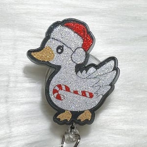 May include: A glittery duck-shaped badge reel with a Santa hat and candy cane design. The duck has a silver body, gold beak and feet, and a black outline. The Santa hat is red with a white trim.