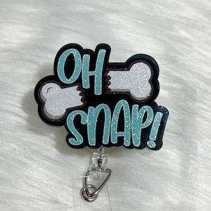 May include: A black and teal blue glitter badge reel with a white bone and the text "OH SNAP!"