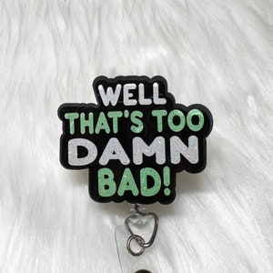 May include: A black and green glitter badge holder with a white and green glitter text that reads "Well that's too damn bad!"