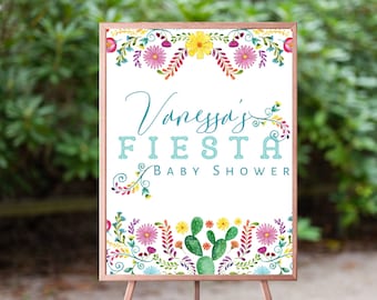 May include: A white sign with a wooden frame and a floral border. The sign says "Vanessa's Fiesta Baby Shower" in blue and white text.
