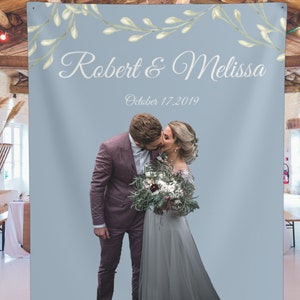 Puede incluir: Un tel&oacute;n de fondo de boda azul claro con una guirnalda floral blanca y el texto "Robert & Melissa" y "Octubre 17, 2019". Una pareja se besa en el centro del tel&oacute;n de fondo.
