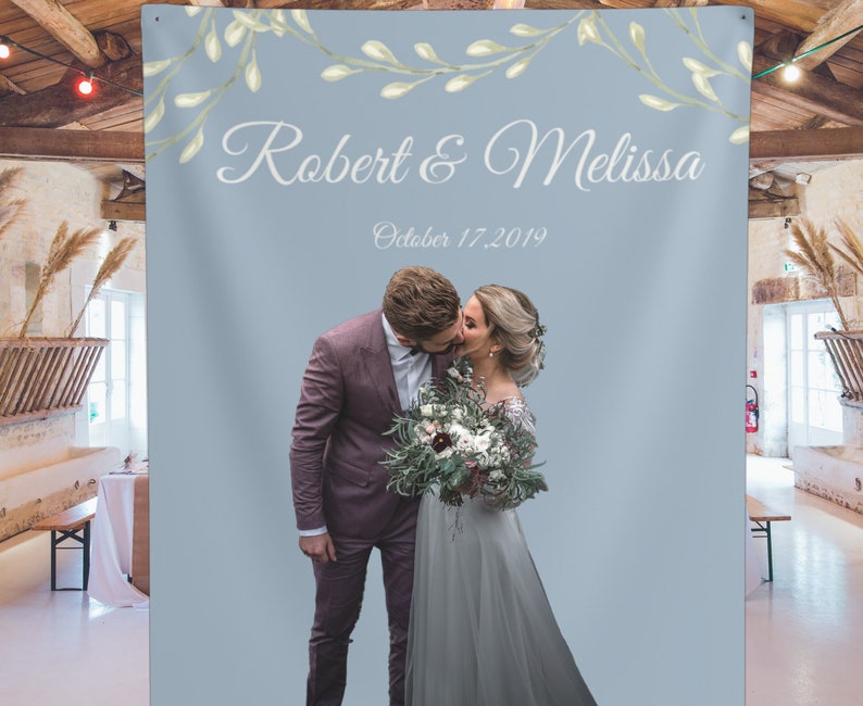 Puede incluir: Un tel&oacute;n de fondo de boda azul claro con una guirnalda floral blanca y el texto "Robert & Melissa" y "Octubre 17, 2019". Una pareja se besa en el centro del tel&oacute;n de fondo.