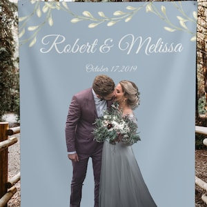 Puede incluir: Un tel&oacute;n de fondo de boda con un fondo azul claro y una guirnalda floral en la parte superior. El texto "Robert & Melissa" est&aacute; escrito en cursiva blanca. La fecha "October 17, 2019" est&aacute; escrita debajo de los nombres. Una foto de una pareja bes&aacute;ndose est&aacute; en la parte inferior central del tel&oacute;n de fondo.