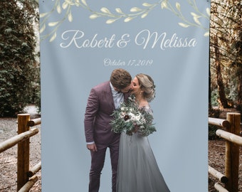 May include: A wedding backdrop with a light blue background and a floral garland at the top. The text "Robert & Melissa" is written in white cursive script. The date "October 17, 2019" is written below the names. A photo of a couple kissing is in the bottom center of the backdrop.