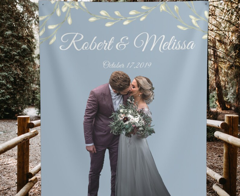 Puede incluir: Un tel&oacute;n de fondo de boda con un fondo azul claro y una guirnalda floral en la parte superior. El texto "Robert & Melissa" est&aacute; escrito en cursiva blanca. La fecha "October 17, 2019" est&aacute; escrita debajo de los nombres. Una foto de una pareja bes&aacute;ndose est&aacute; en la parte inferior central del tel&oacute;n de fondo.