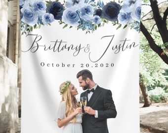 May include: A white wedding backdrop with a blue floral garland at the top. The text "Brittany & Justin" and "October 20, 2020" is printed below the garland. A photo of a couple is in the bottom half of the backdrop.