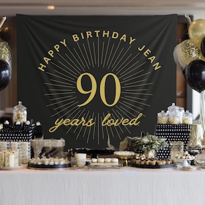 May include: A festive birthday party setup with a black banner reading "Happy Birthday Jean 90 years loved" in gold. Balloons in black and gold, along with desserts and decorations, adorn the table.