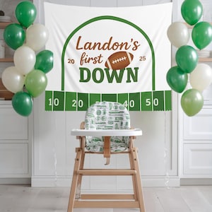 May include: A high chair with a green and white patterned seat is set up in front of a banner that reads "Landon's first DOWN" with a football graphic. Green and white balloons are tied to the sides. The banner has a football field design.