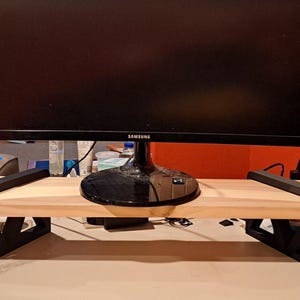 May include: A monitor stand featuring a rectangular light-colored wooden shelf supported by two black, triangular-patterned legs. A black Samsung computer monitor is positioned on top of the wooden surface, centered between the two black supports.