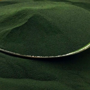 Puede incluir: Primer plano de un polvo verde oscuro, posiblemente un suplemento o ingrediente alimenticio, que llena un cuenco con borde plateado. El polvo tiene una textura fina y granulada y un color profundo y rico, lo que sugiere un producto natural.