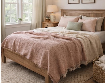 May include: A wooden bed frame featuring a textured, dusty rose-colored throw blanket draped over the foot. The bedding includes light pink and cream pillows, a small sage green accent pillow, and a woven cream-colored area rug on the floor.