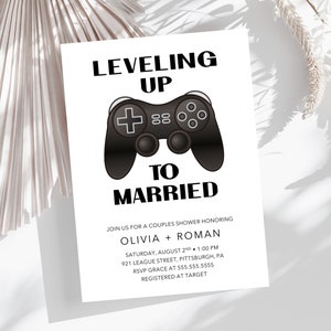 May include: A white card with black text that reads "Leveling Up to Married."  The card features a black and white illustration of a video game controller.  The text also includes the names Olivia and Roman, the date and time of the event, the location, and RSVP information.