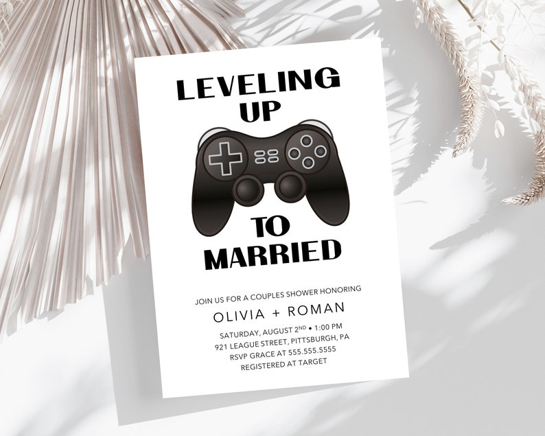 May include: A white card with black text that reads "Leveling Up to Married."  The card features a black and white illustration of a video game controller.  The text also includes the names Olivia and Roman, the date and time of the event, the location, and RSVP information.