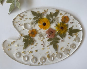 May include: A white oval-shaped resin tray with a thumb hole and eight circular wells. The tray is decorated with pressed flowers and gold leaf.