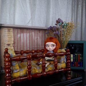 May include: A miniature wooden bed with a yellow plaid cushion. A doll with red hair sits on the bed, holding a book. The bed is made of dark wood and has a decorative headboard and footboard. A small bouquet of dried flowers sits nearby.