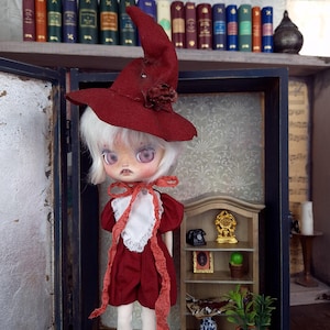 May include: A doll wearing a red witch hat and a red dress with white lace trim. The doll is standing in front of a miniature bookshelf with a clock, a phone, and a potted plant.