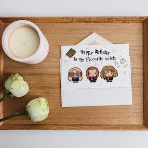 May include: A white card with a glittery background and the text "Happy Birthday to my favourite witch". Three cartoon witches are depicted on the card, with a book, a potion bottle, and a heart.