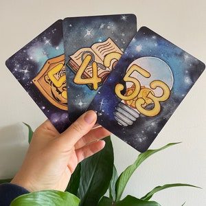 May include: Three cards with a space-themed background. Each card features a different number, 4, 5, and 3, in a yellow, cartoon font. The number 4 is on a card with an open book, the number 5 is on a card with a lightbulb, and the number 3 is on a card with a shield.