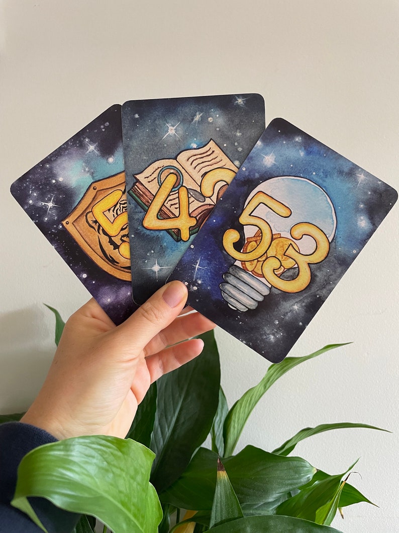 May include: Three cards with a space-themed background. Each card features a different number, 4, 5, and 3, in a yellow, cartoon font. The number 4 is on a card with an open book, the number 5 is on a card with a lightbulb, and the number 3 is on a card with a shield.