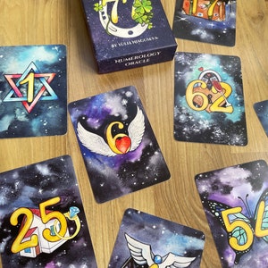 May include: A set of tarot cards with a galaxy background and gold numbers. The cards feature various symbols, including a horseshoe, a house, a butterfly, a heart, a scale, and a suitcase. The cards are part of a numerology oracle deck.