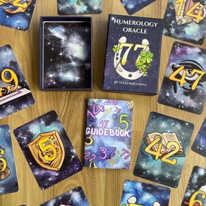 May include: A numerology oracle deck with a guidebook. The cards feature a galaxy background with numbers and symbols. The guidebook has a galaxy pattern with numbers printed on the cover.