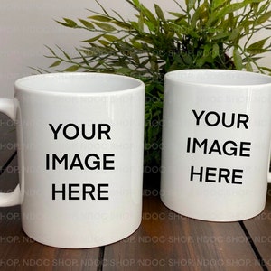 Puede incluir: Dos tazas de cerámica blanca con texto negro que dice "YOUR IMAGE HERE". Las tazas están sobre una superficie de madera con plantas verdes frondosas en el fondo.