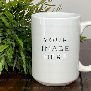 Puede incluir: Taza de cerámica blanca con un texto negro superpuesto que dice "YOUR IMAGE HERE". La taza está sobre una superficie de madera junto a una planta verde en maceta.