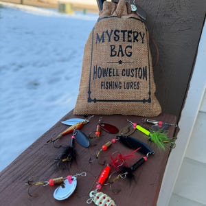 May include: A burlap bag labeled "MYSTERY BAG HOWELL CUSTOM FISHING LURES" sits on a wooden surface, surrounded by various fishing lures. The lures feature metallic blades, colorful beads, and feathered accents, in a variety of shapes and sizes.