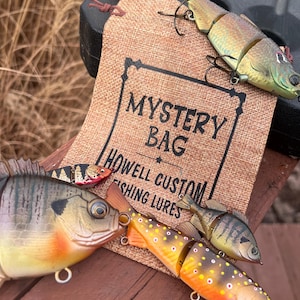May include: Four different fishing lures in a mystery bag. The bag is made of burlap and has the text "Mystery Bag Howell Custom Fishing Lures" printed on it. The lures are all different colors and designs, and they are all made of plastic.