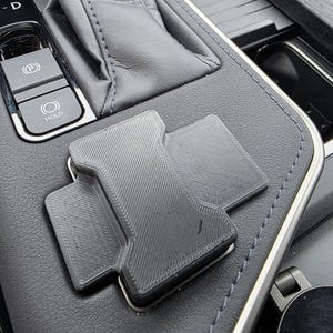 May include: A black, 3D-printed car phone mount, designed to fit a vehicle's console. The mount is rectangular with a textured surface and is positioned on a black leather surface with silver trim and blue stitching.