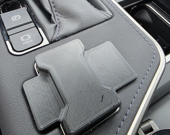 May include: A black, 3D-printed car phone mount, designed to fit a vehicle's console. The mount is rectangular with a textured surface and is positioned on a black leather surface with silver trim and blue stitching.
