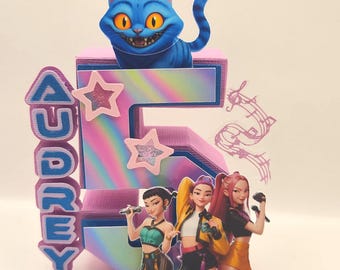 May include: A colorful cake topper featuring a blue Cheshire Cat figure atop a large number 5. The number is decorated with a rainbow gradient and pink stars. The word "AUDREY" is spelled out vertically. Three cartoon figures are at the base.