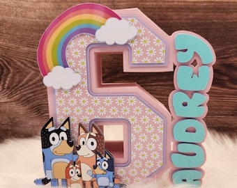 May include: A pink number six cake topper with a rainbow and clouds on top. The number six is decorated with a daisy pattern. The cake topper also includes a blue and green name tag that says "Audrey". There are four cartoon dogs in front of the number six.