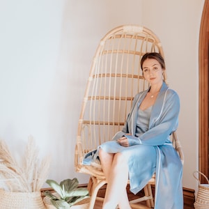 May include: A person wearing a light blue satin robe with white piping sits in a woven wicker chair. The chair is in a room with a light wood floor and white walls.