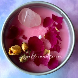 Könnte beinhalten: Eine rosa Kerze mit getrockneten Rosenblüten und einem Rosenquarzkristall. Die Kerze befindet sich in einer silbernen Dose mit dem Text "spell candle co."