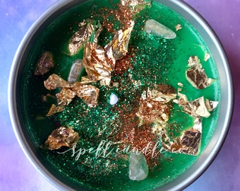 May include: A green candle with gold flakes, glitter, and clear crystals. The candle is in a silver tin with the text "spellcandleco" on the side.