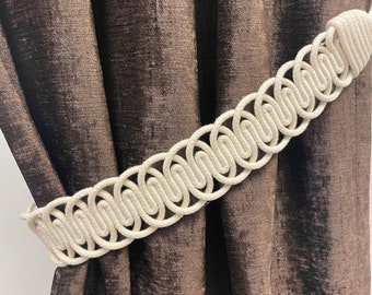 May include: A white rope curtain tieback with a decorative woven design. The tieback is holding back brown velvet curtains.