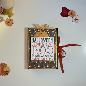Peut inclure: Un album de scrapbooking sur le thème d'Halloween noir et or avec une couverture à motifs noir et blanc. La couverture porte le texte "HALLOWEEN OCTOBER 31ST BOO trick or treat HAPPY HAUNTING".